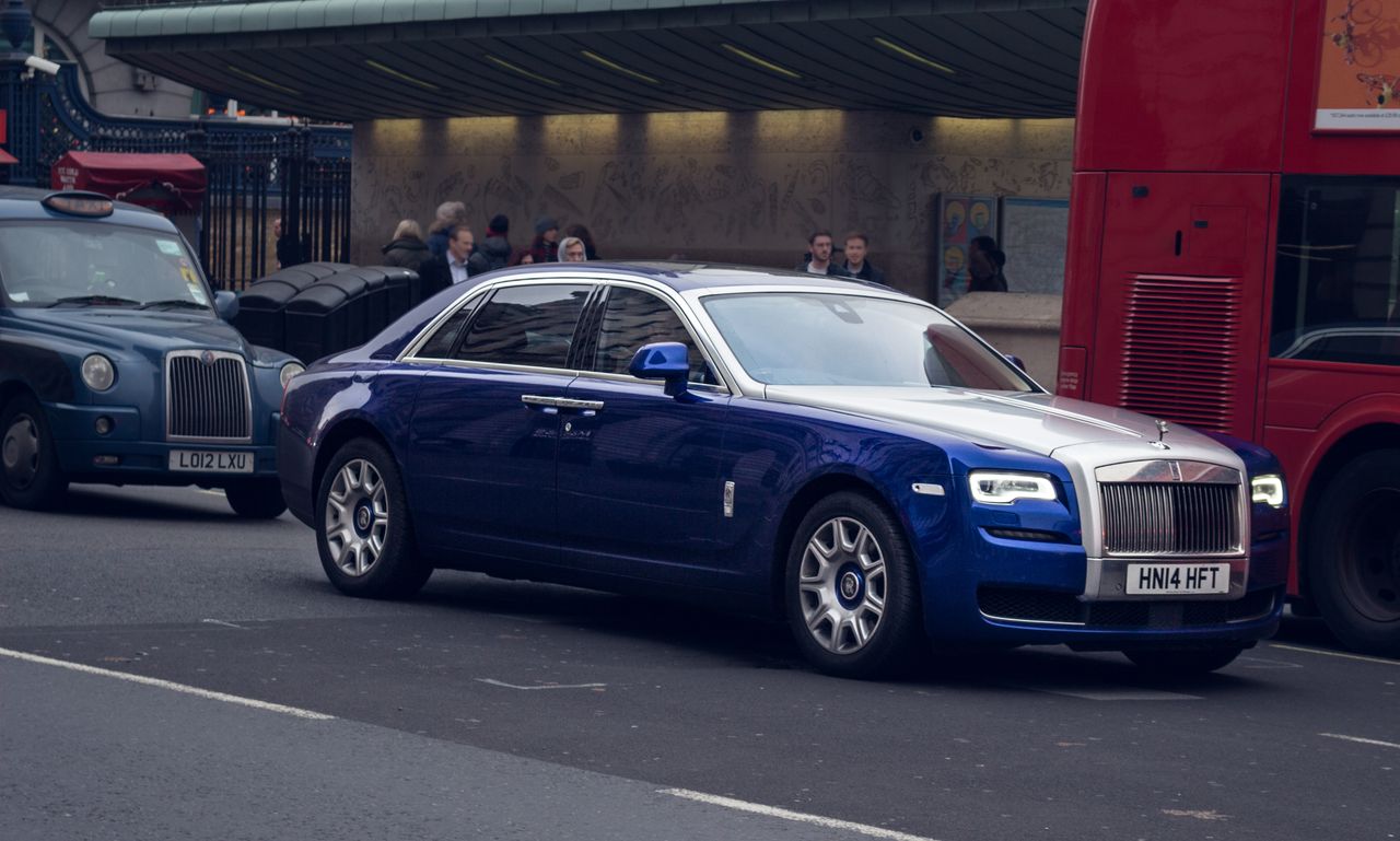 A blue luxury car driving on a busy street in London, surrounded by iconic vehicles.